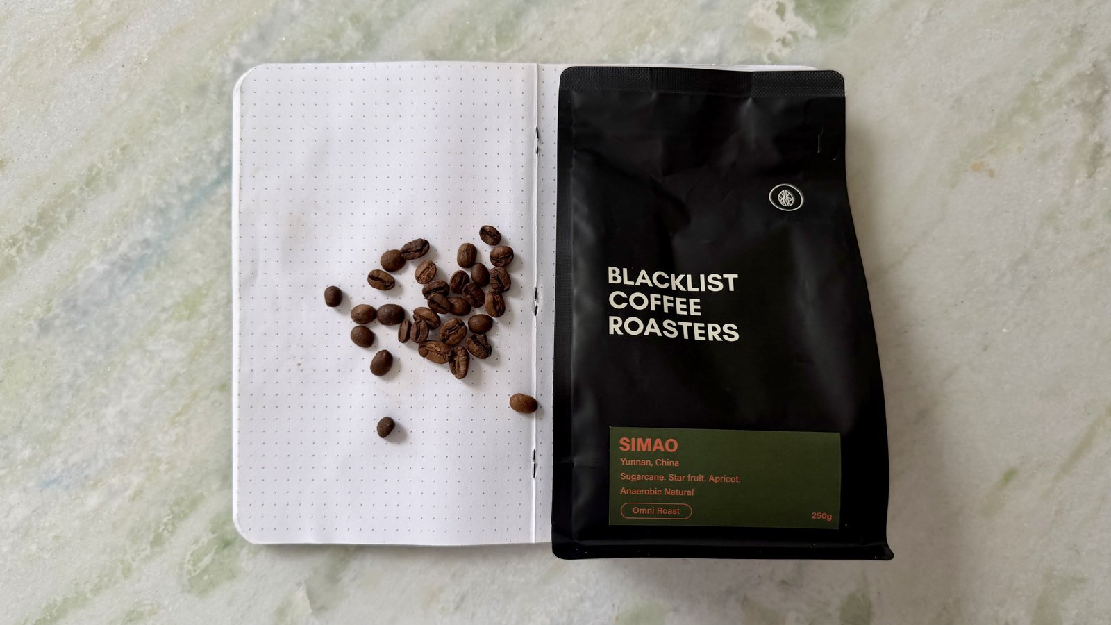 Close-up of roasted coffee beans on an open notebook with a black coffee bag from Blacklist Coffee Roasters in the background, showcasing high-quality specialty coffee.
