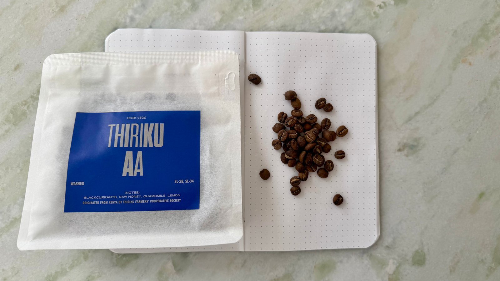 Coffee beans and a notebook with a dotted page, featuring a blue label reading "THIRIKU AA," arranged on a light marble background.