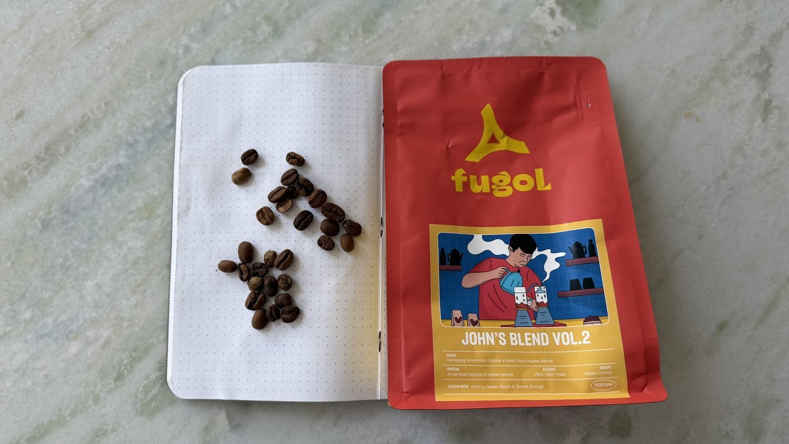 Coffee beans arranged on a dotted notebook page next to a red FUGOL coffee bag featuring a colourful illustration.