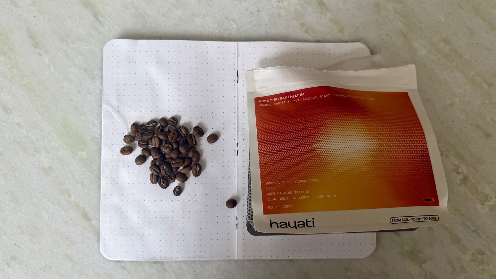 Close-up of coffee beans on a white notebook with a red and orange packet from Hayati on a light surface.
