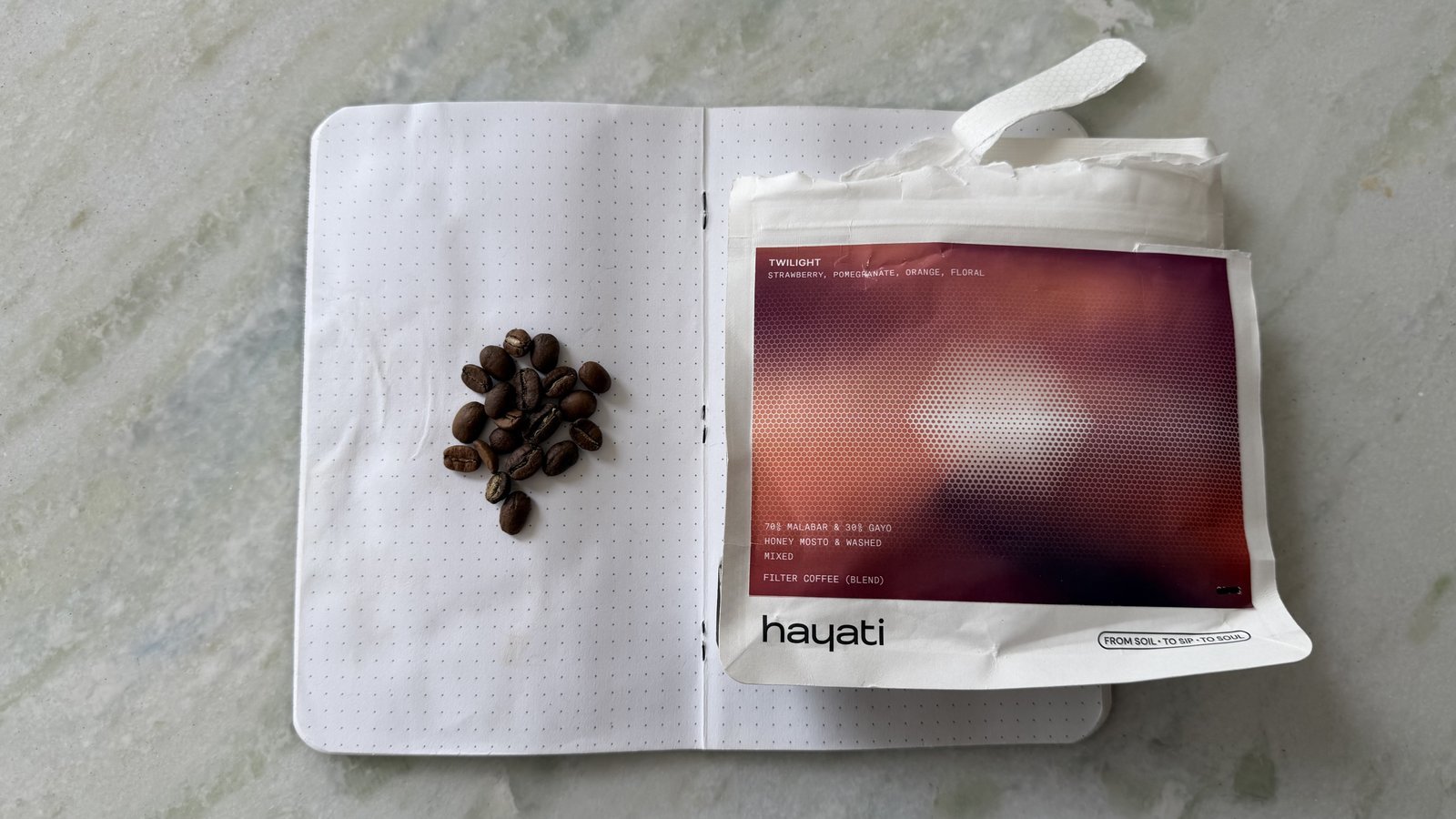 Close-up of coffee beans on an open notebook with a coffee package on a light grey surface.